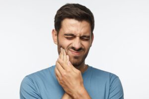 Portrait of young man isolated on gray background, her face distorted and hands pressed to her cheek in pain tmj treatment dentist in Manhattan New York
